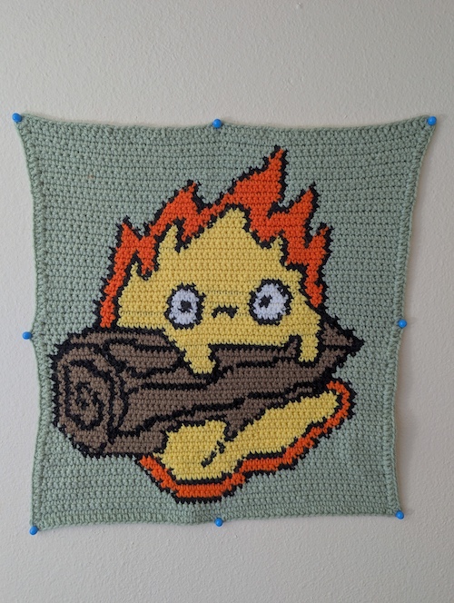 A crocheted wall tapestry depicting Calcifer resting on a log