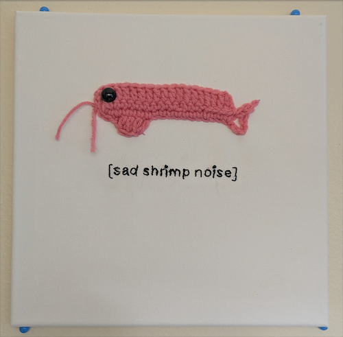 A small pink crocheted shrimp mounted on white canvas above the words sad shrimp noise