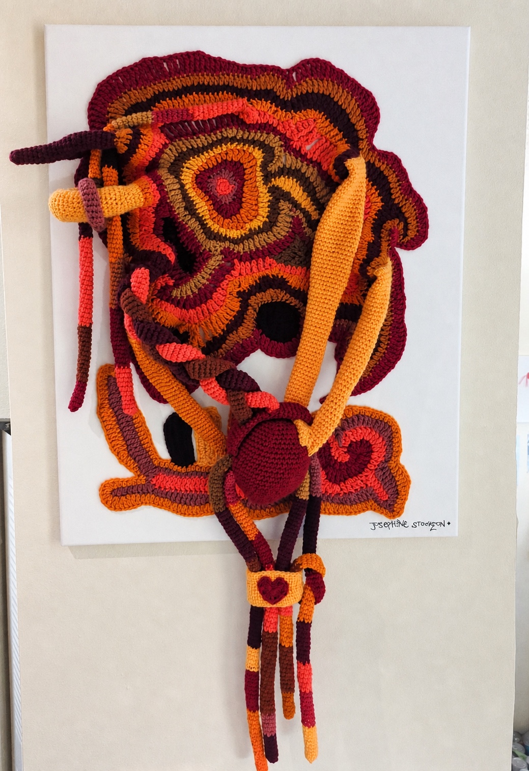 A warm-toned freeform fiber artwork with layered crochet forms and a dangling anatomical heart