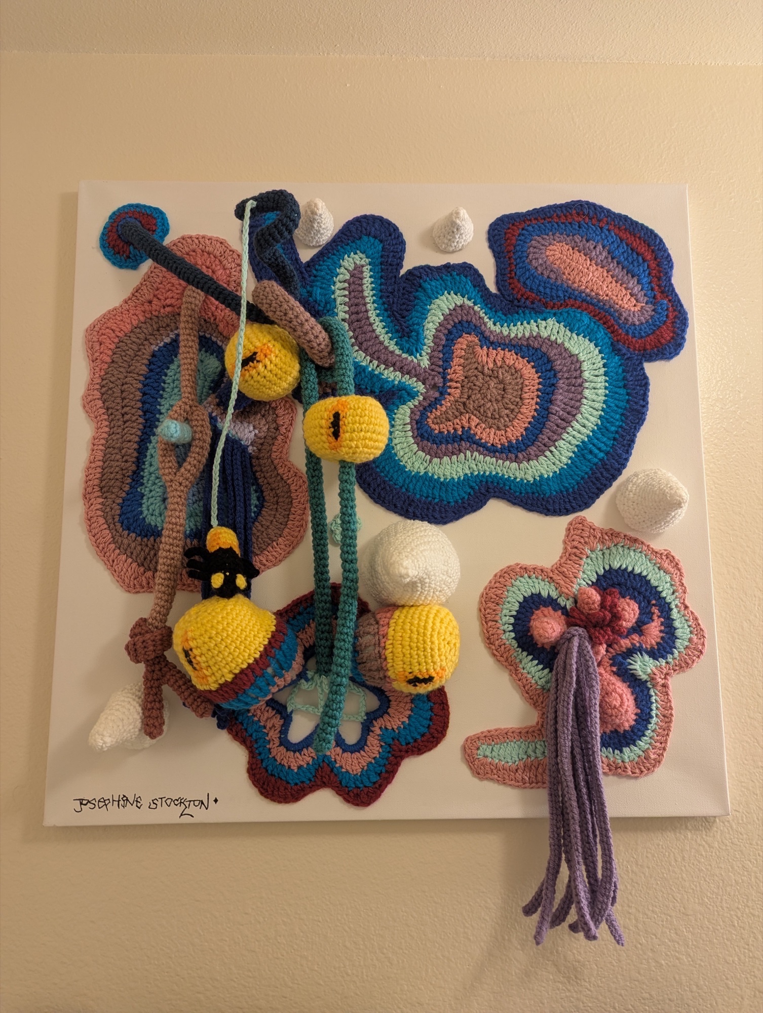 A blue, pink, and purple freeform fiber artwork with yellow eyes and dangling swamp-like creatures